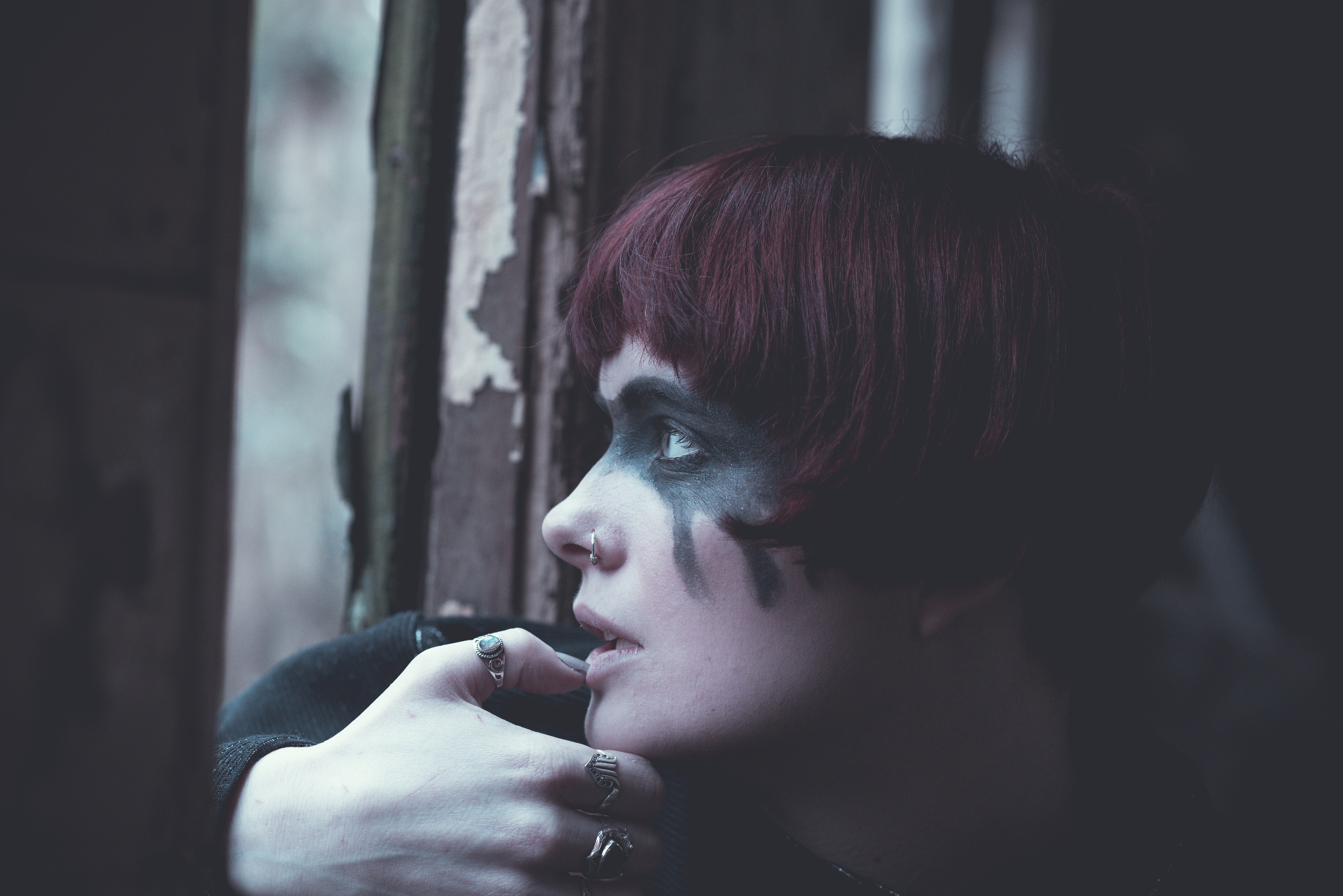 Foto de uma garota de cabelos curtos e ruivos, com uma maquiagem gótica pesada. Está sentada próxima a uma janela, de um local que parece ser um cemitério.